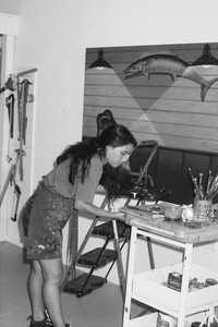 Black-and-white photograph of Julie Curtiss in her studio leaning over a stand of painting materials with a painting featuring a fish hanging on the wall