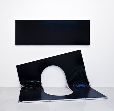 Installation featuring a black rectangular painting hanging on a wall and a second rectangular painting with an oval cut-out in the center folded in half resting on the floor below