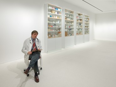 Realistic bronze sculpture of a medical doctor seated in an office chair with a stethoscope and clipboard in front of a four-part medicine cabinet with shelves stocked with drug packages