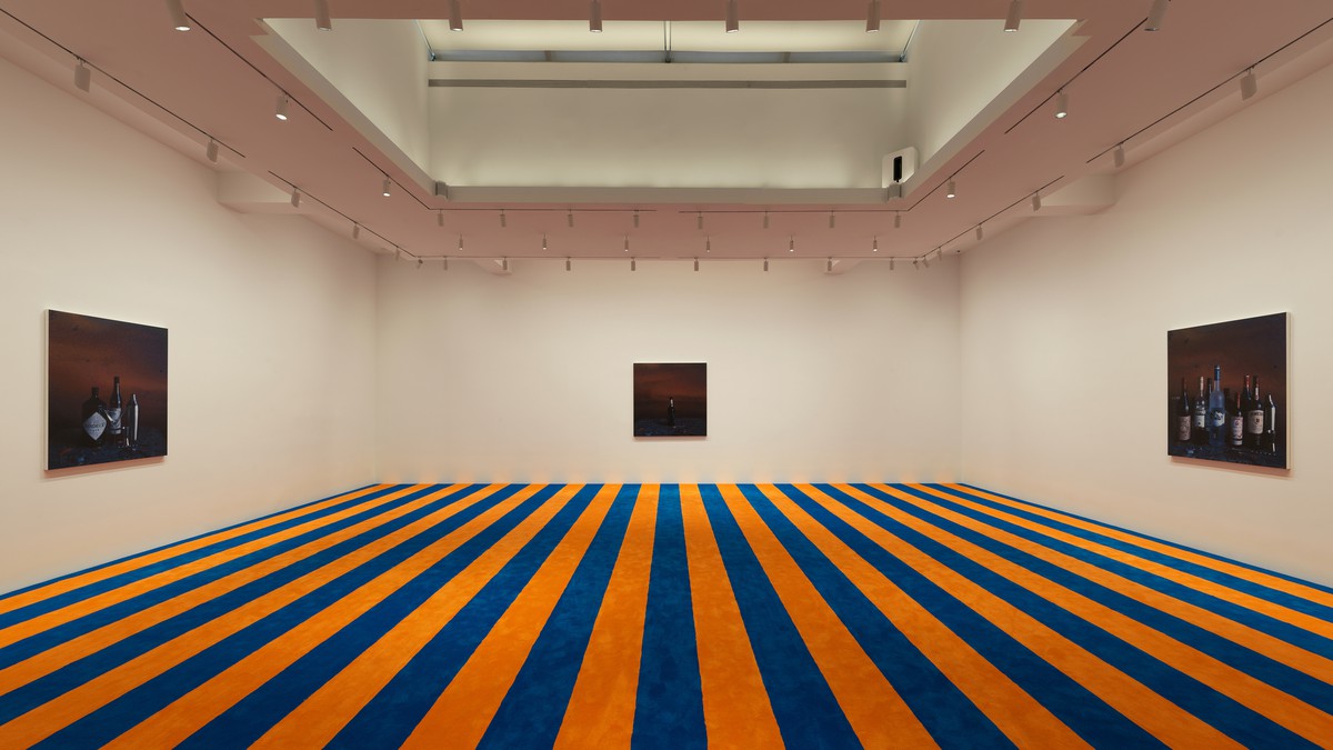 Installation of three paintings of various alcohol bottles in a gallery with a blue and orange striped carpet