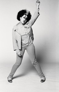 Black and white portrait of Poly Styrene with one hand in the air and knees knocked in