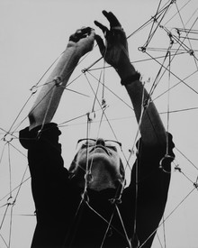 Black and white photo of Gego looking up and holding sculptural wires while installing Reticulárea, Museo de Bellas Artes de Caracas, 1969.