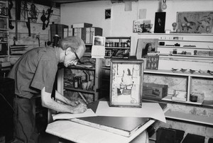 Black-and-white photograph of a man in an art studio bent over a table with pen to paper
