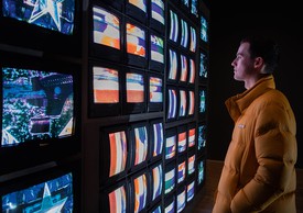 person in yellow puffer jackets stands in front of a plethora of tv screens