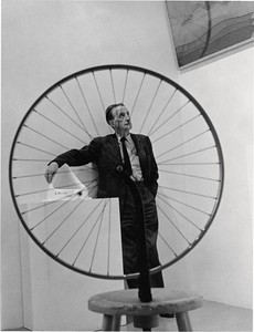 Black and white image of Marcel Duchamp standing behind his work "Bicycle Wheel"