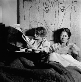 Black-and-white photograph of two women having breakfast in bed and reading newspapers with a tapestry of an abstracted face hanging on the wall behind them