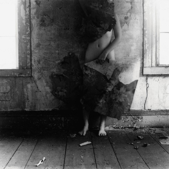 Self portrait of Francesca Woodman, she stands against a wall holding pieces of ripped wallpaper in front of her face and legs