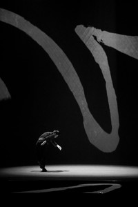 black-and-white photograph of a man dancing in a spotlight with a projection of curving brushstrokes on the stage background