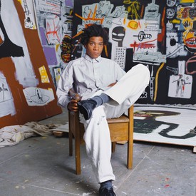 Portrait of Jean-Michel Basquiat sitting inside his studio and in front of his paintings
