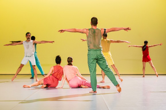 Multiples dancers in bright costumes against a yellow backdrop. Five have their backs to the camera with their arms stretched out and two are sitting center stage.