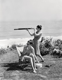 Portrait of twins Frances McLaughlin-Gill and Kathryn Abbe in front of a beach, one of them sits in a lawn chair and the other stands behind looking out of a spyglass