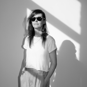 Black-and-white image of Lucy Raven sitting in a field with a sunglasses and a bandana over her hair next to a table with a bottle of wine.