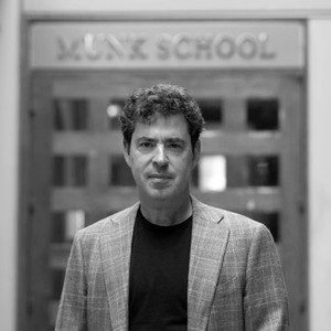 Portrait of a man with curly dark hair and light skin in front of a door reading "Munk School" wearing a blazer over a dark shirt
