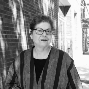 Black-and-white portrait of Donna De Salvo outdoors in front of a brick building. She has short hair, glasses, dark lipstick, and a top with vertical stripes.
