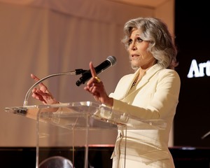 Jane Fonda wearing a white suit and speaking at a podium at the Art for a Safe and Healthy California benefit launch