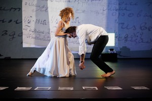 A woman wearing a white dress with her leg outstretched behind her holds a mans head in her hands; they are facing each other in profile. Behind them is a projection of archival letters and printed photographs sit at the front of the stage.