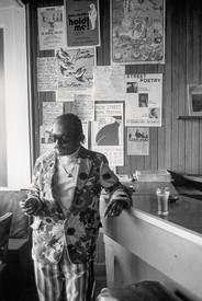A black-and-white photograph of the poet Bob Kaufman leaning against a counter wearing a floral jacket, striped pants, and sunglasses.