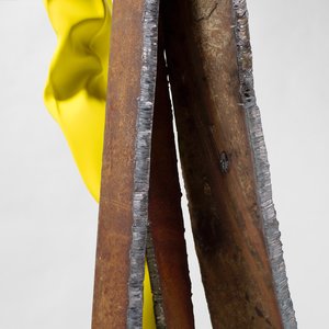 Detail of an approximately ten-foot-tall slender, vertically oriented abstract sculpture comprising a crumpled yellow stainless-steel tube and raw mild steel
