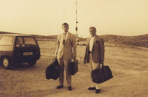 Two men in suits holding onto luggage and briefcases in front of a parked car
