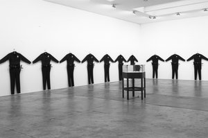 Gallery with ten oversize police uniform hanging in a visual cordon on the wall and a sculpture in the center of the room
