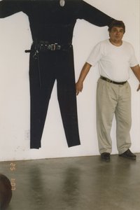 Chris Burden standing with his back against a wall with an oversize uniform hanging on it