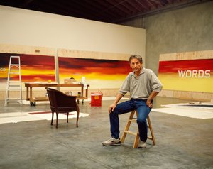 Image of Ed Ruscha sitting on a stool in his studio in front of three paintings
